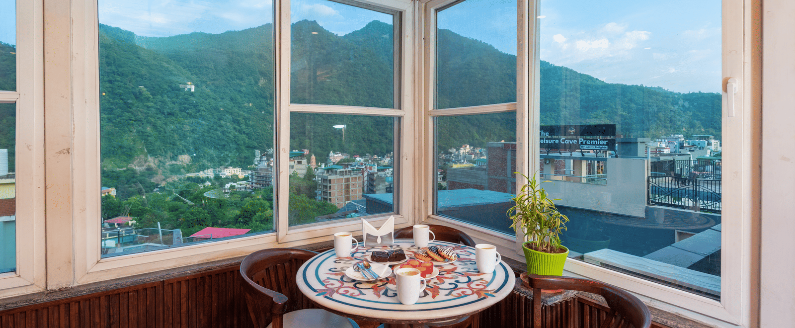 A cafe dining area with a round table set for a meal, including drinks, a cake, and pastries. The room has large windows offering a panoramic view of lush green hills and the town below, with a wooden-paneled ceiling and walls. This is a dining area at Reg