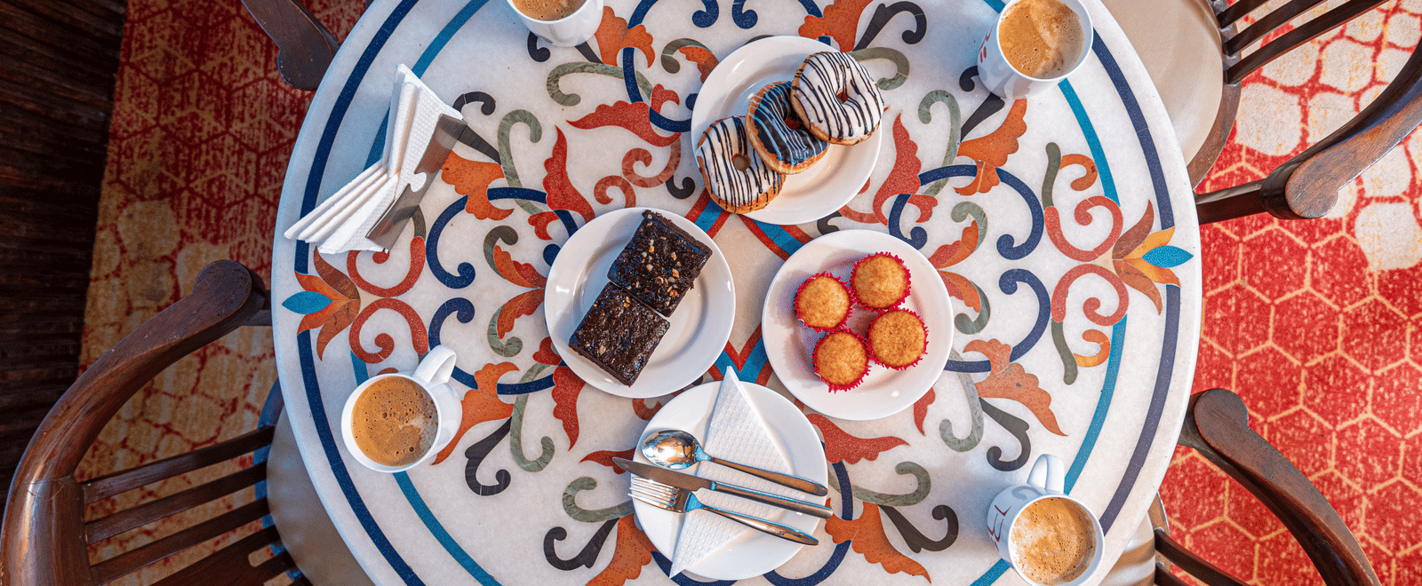 An overhead shot of a circular table set with a variety of pastries, brownies, and four cups of coffee. The table has a colorful, intricate floral and geometric pattern, with four chairs arranged around it. This is a food shot from the cafe at DLS Hotels O