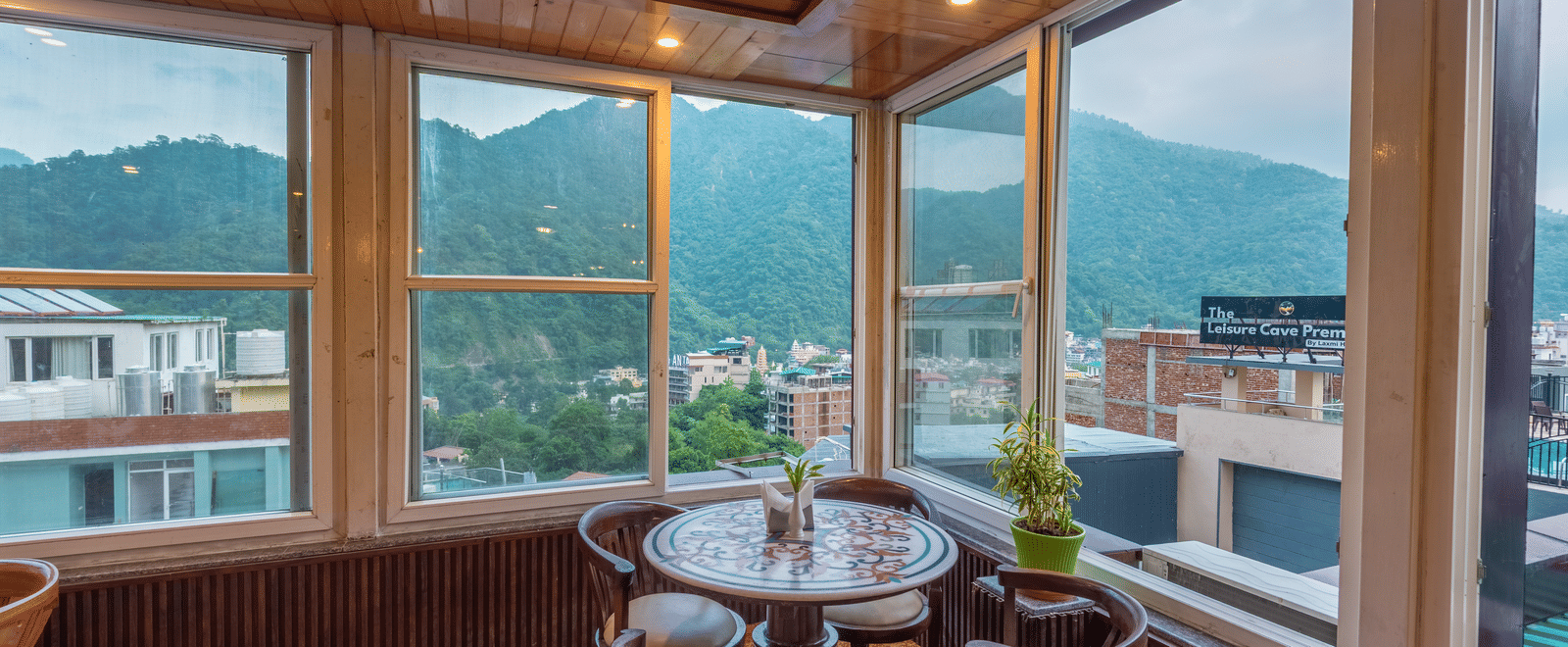 A cafe dining area at dusk with a round table set for four. The room has large windows on two sides, showcasing a view of the mountains and the town. The interior has a warm glow from the lights, highlighting the wooden ceiling and furniture. This is a din