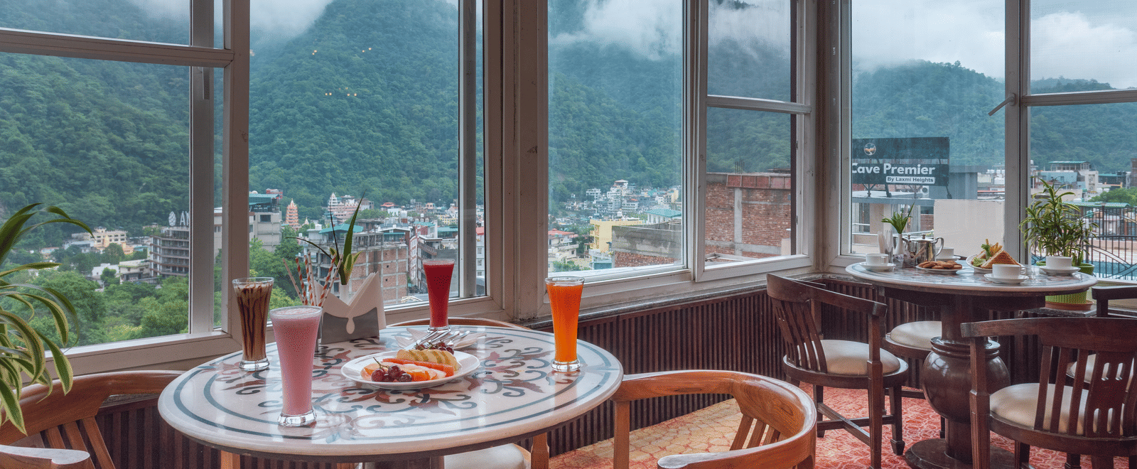 A cafe with a vibrant, red patterned carpet and wooden furniture. The room has large windows that look out onto misty green mountains and the city below. Tables are set with various drinks and food, including a fruit platter and sandwiches. This is the caf