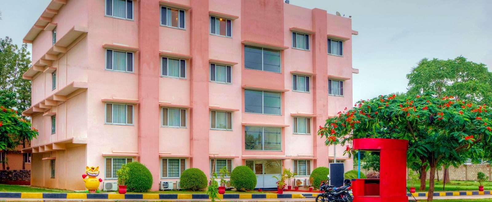A vibrant exterior shot of a pink hotel building with greenery and a red security booth in the foreground at Greens Inn - Inviting Hotel in Hyderabad.