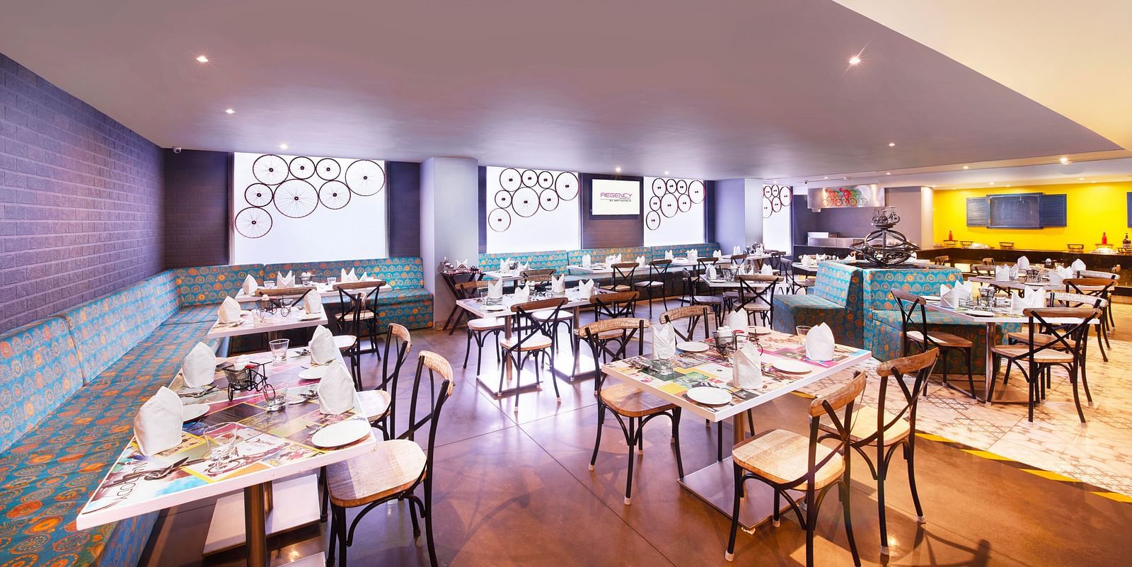 A brightly lit restaurant with chairs, a long bar counter, and a colorful wall mural | Regency Tirunelveli By GRT Hotels