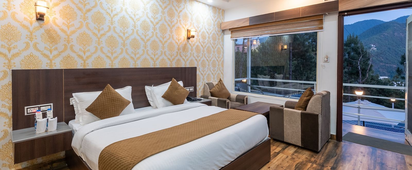 Elegant hotel room with a large double bed, warm wooden flooring, patterned wallpaper, a sitting area by a large window, and a balcony offering scenic views of the mountains and trees - DLS Nature Trinket Resort, Dalhousie.