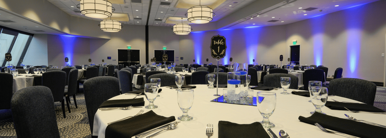 A wide view of a banquet hall set up with large, round tables, white linens, black napkins, and modern chandeliers. 