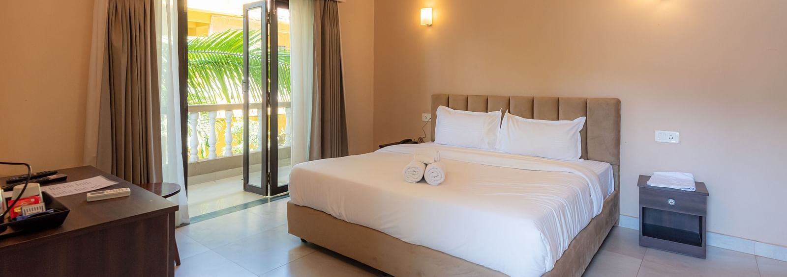Spacious room with a large bed, wooden bedside table, wall-mounted lamp above the bed, and a glass doorway to the balcony at De Soul Santé, Morjim.