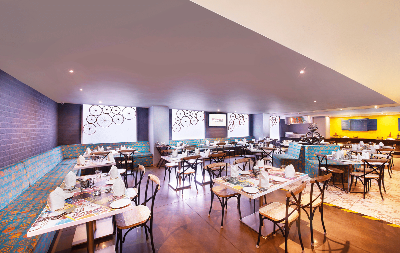 A brightly lit restaurant with chairs, a long bar counter, and a colorful wall mural | Regency Tirunelveli By GRT Hotels
