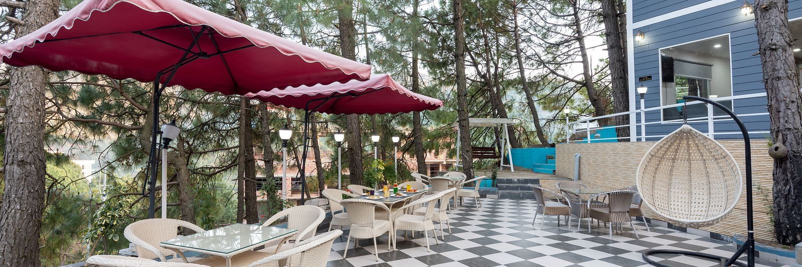 Charming outdoor seating area with checkered tile flooring, glass-top tables, and white wicker chairs, set beneath red canopies among tall pine trees next to a modern blue building with balconies - DLS Nature Trinket Resort, Dalhousie.