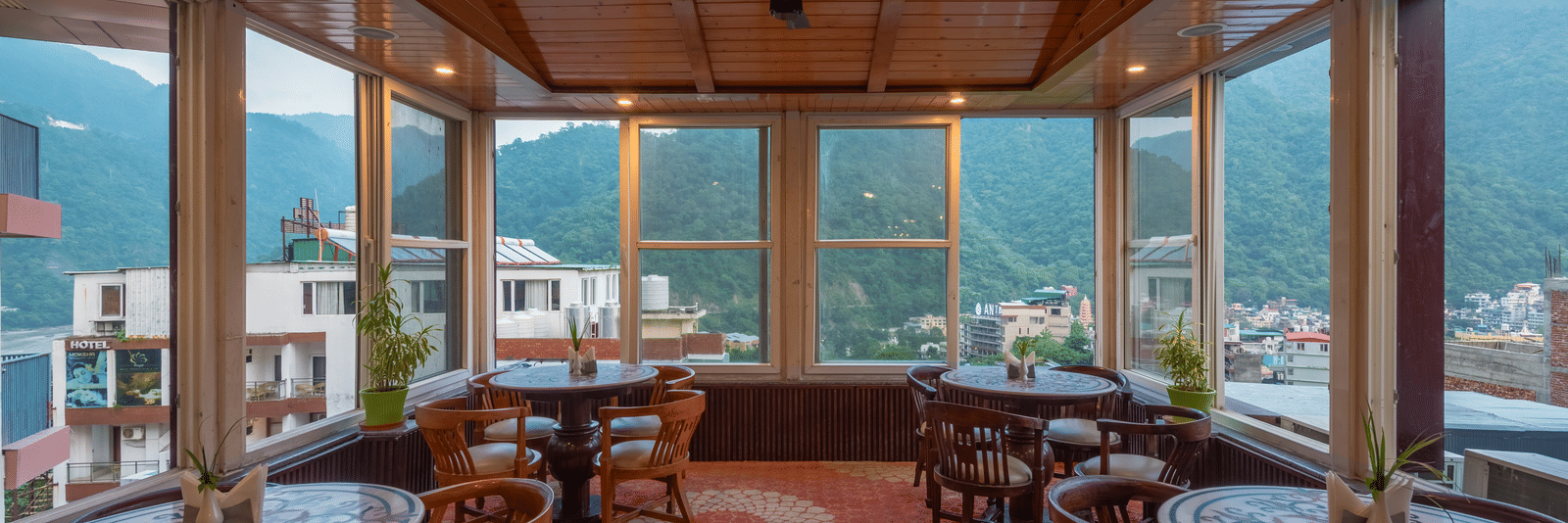 A bright, sunlit cafe with multiple tables, each set with food and drinks. The space is surrounded by large windows, offering an expansive view of green, forested mountains and buildings in the valley. This is the cafe at DLS Hotels On The Ganges.