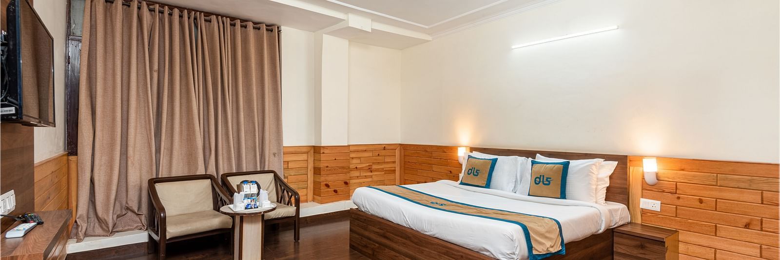 A well lit hotel room with a wooden floor, a large bed, two armchairs, a small table, and a ceiling fan | DLS Anupam Resort, Dharamshala