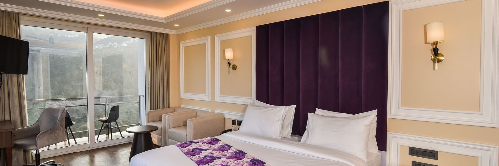 A vibrant bedroom with a large bed featuring purple accents and a bright atmosphere. The room includes large windows that provide scenic views of the hills outside.