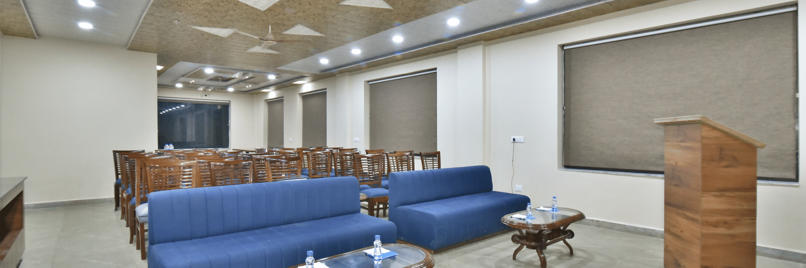 Conference hall at DLS Hotels with wooden chairs, blue sofas, podium, and ceiling fans under warm lighting.