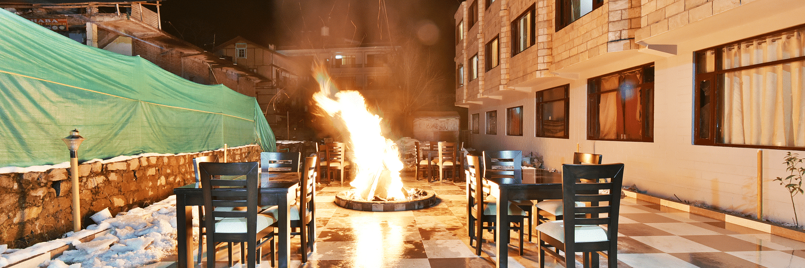 The image shows an outdoor dining area at night with a large bonfire in the centre, surrounded by tables and chairs | DLS Kapoor Resort Manali