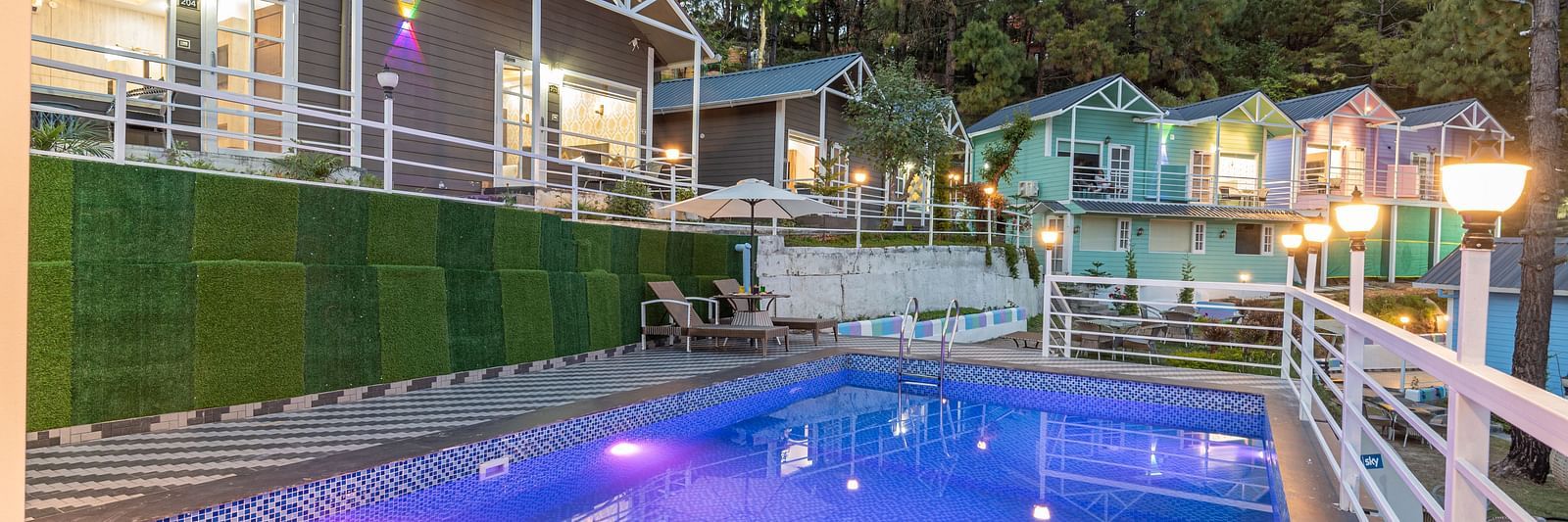 Evening view of a lit-up swimming pool with purple and white underwater lights, surrounded by a safety railing, colourful cottages, and pine trees in a hillside resort setting - DLS Nature Trinket Resort, Dalhousie.