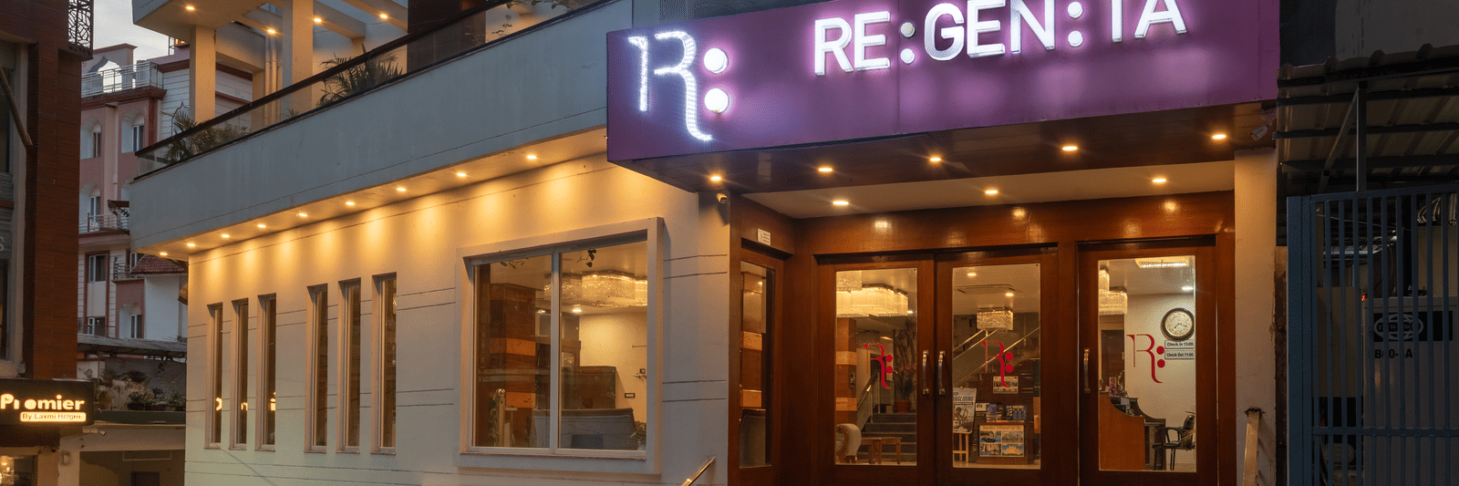 An eye-level view of the entrance to the DLS Hotels On The Ganges, in the evening. The hotel's brightly lit RE:GEN:TA sign is visible, along with the glass-paned doors of the lobby. The exterior walls are a mix of white and dark paneling, with warm light