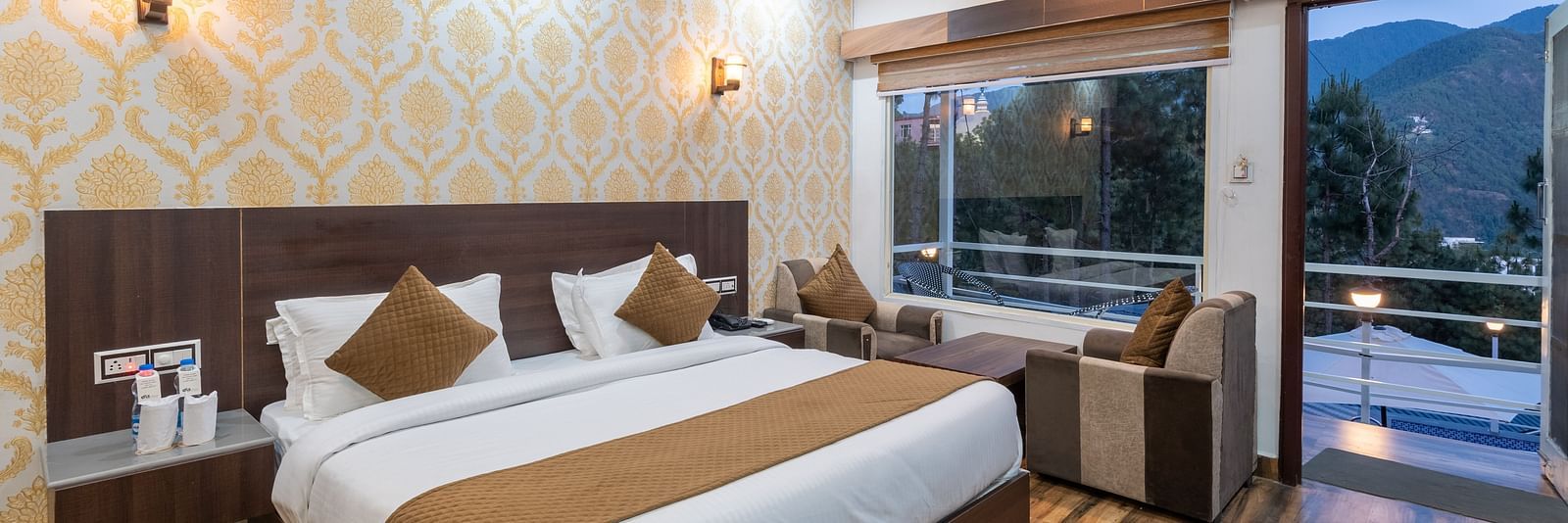 Elegant hotel room with a large double bed, warm wooden flooring, patterned wallpaper, a sitting area by a large window, and a balcony offering scenic views of the mountains and trees - DLS Nature Trinket Resort, Dalhousie.