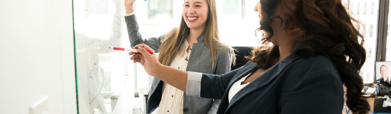Two women are standing in front of a whiteboard, one is writing at the board with a marker, and the other is looking on.