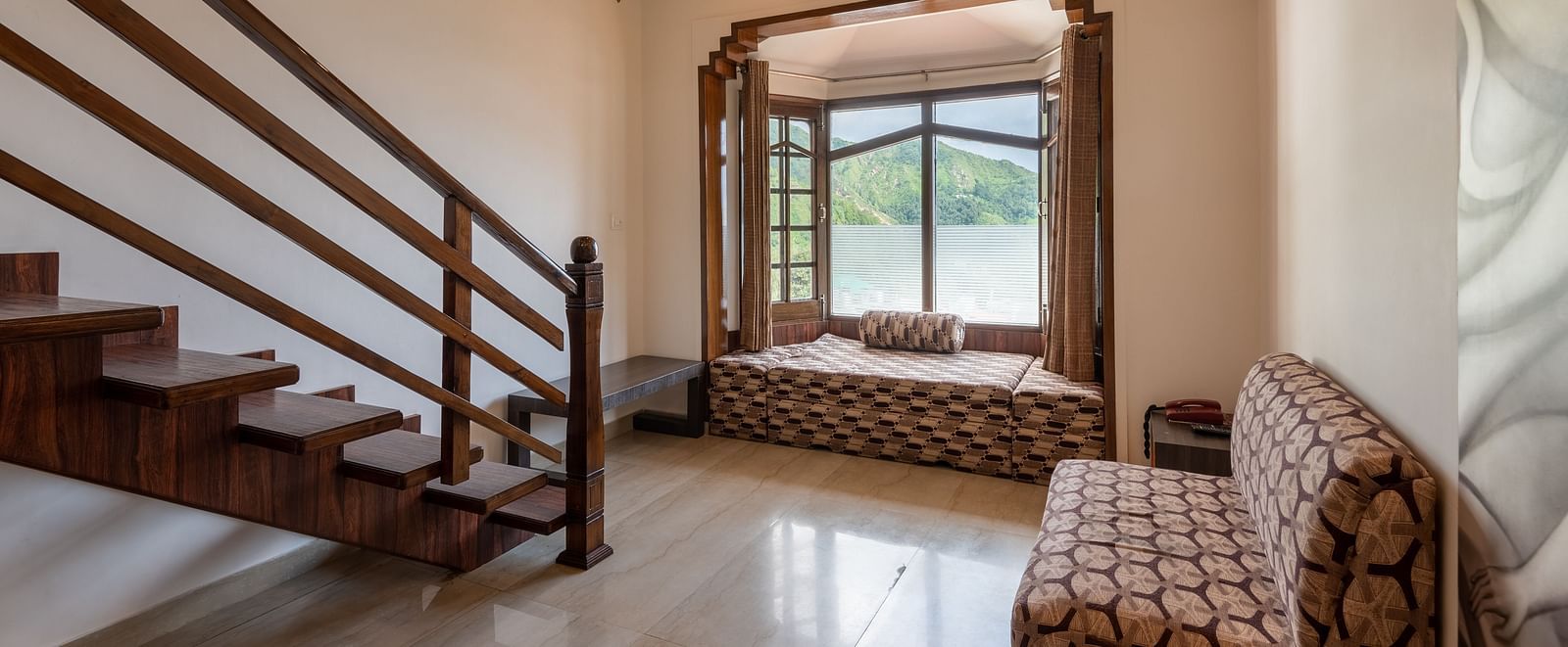 A room with wooden stairs, a seating area by a large window offering a mountain view, and a comfortable couch - DLS Meghavan Resort, Dharamshala