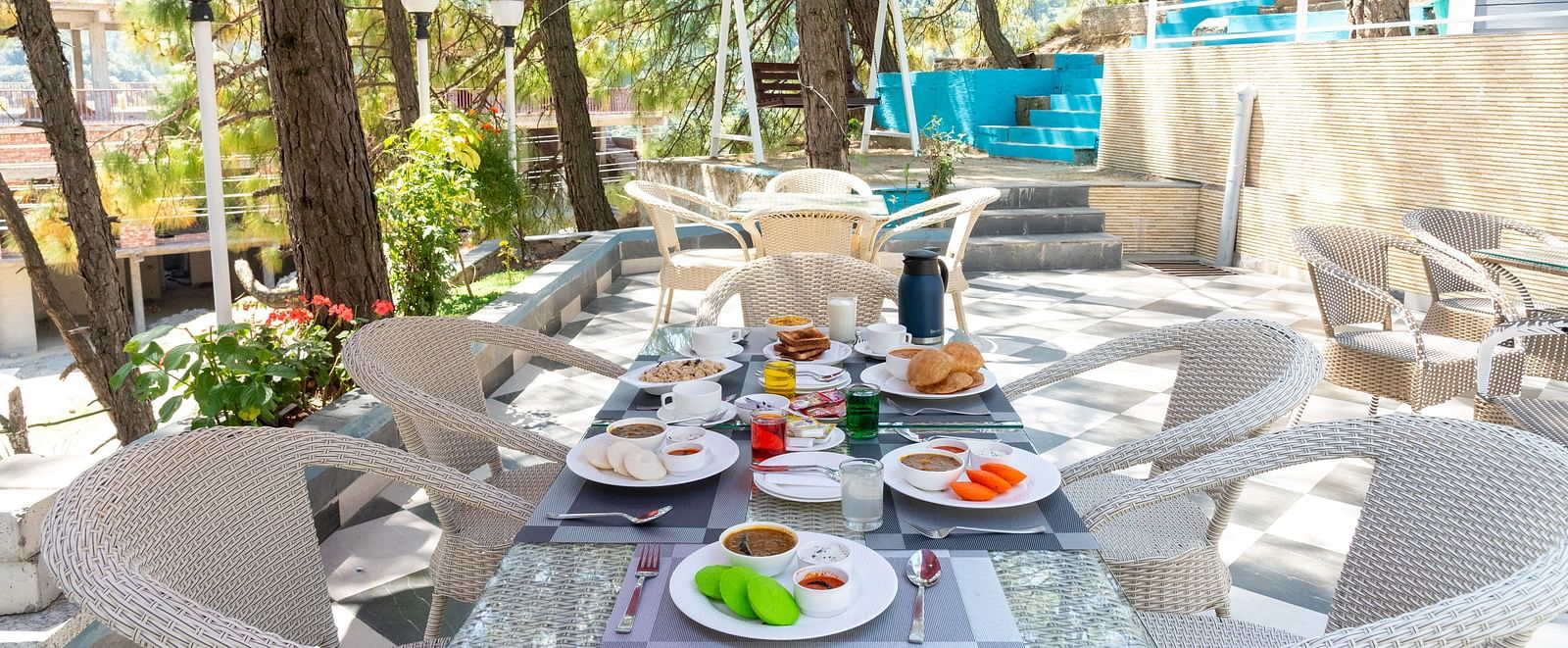 Spacious outdoor dining area set for breakfast, featuring a glass-top table with assorted dishes, surrounded by white chairs under a red canopy, nestled among tall trees and natural greenery - DLS Nature Trinket Resort, Dalhousie.
