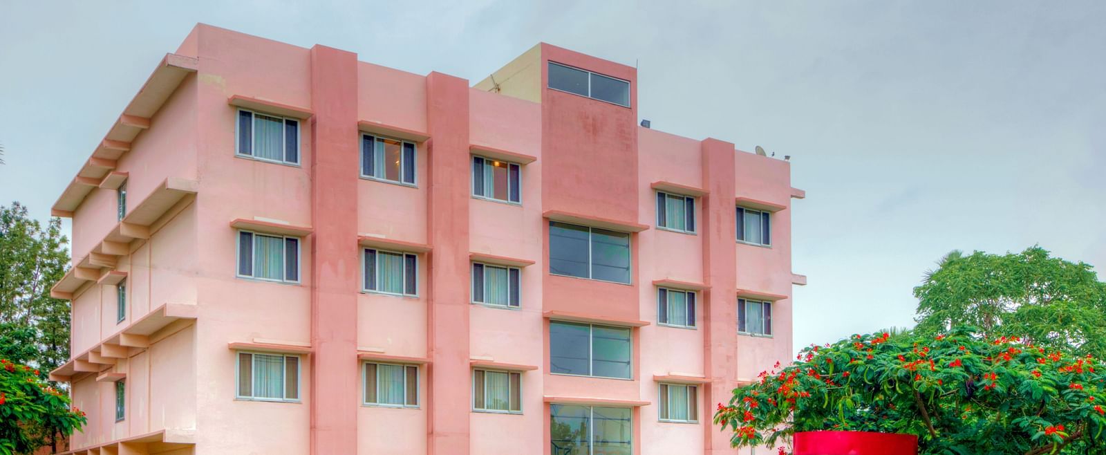 A vibrant exterior shot of a pink hotel building with greenery and a red security booth in the foreground at Greens Inn - Inviting Hotel in Hyderabad.