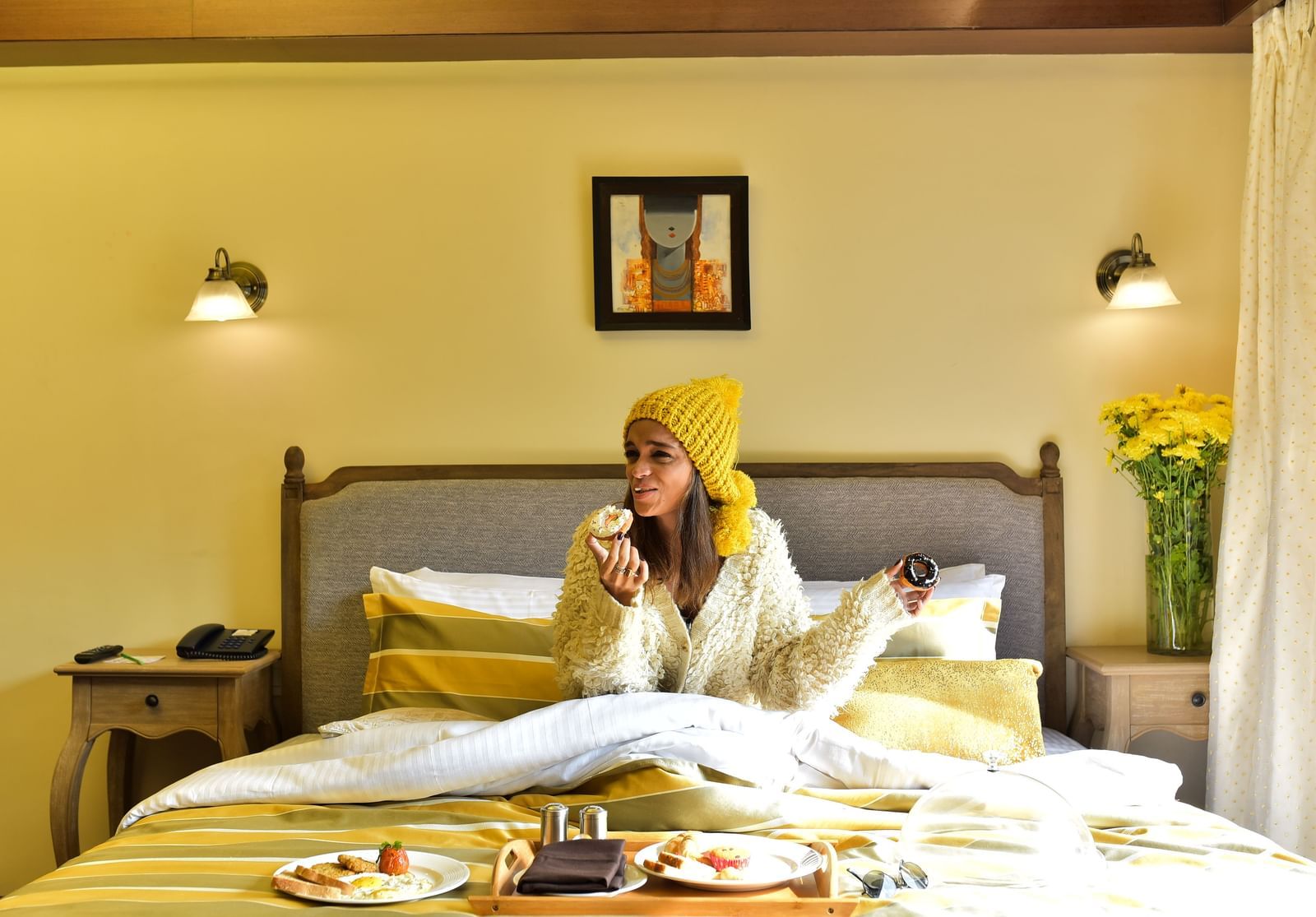 A woman in a yellow beanie and white cardigan enjoys breakfast in bed at the Polo Hotel Shillong.