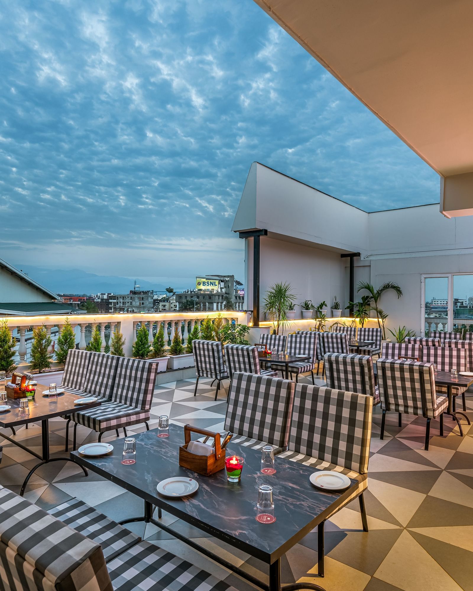 An outdoor rooftop dining area with tables and chairs, set beneath a dramatic cloudy sky. It features a patterned floor, decorative plants, and a partial view of the city  - Lords Eco Inn Dehradun.- 