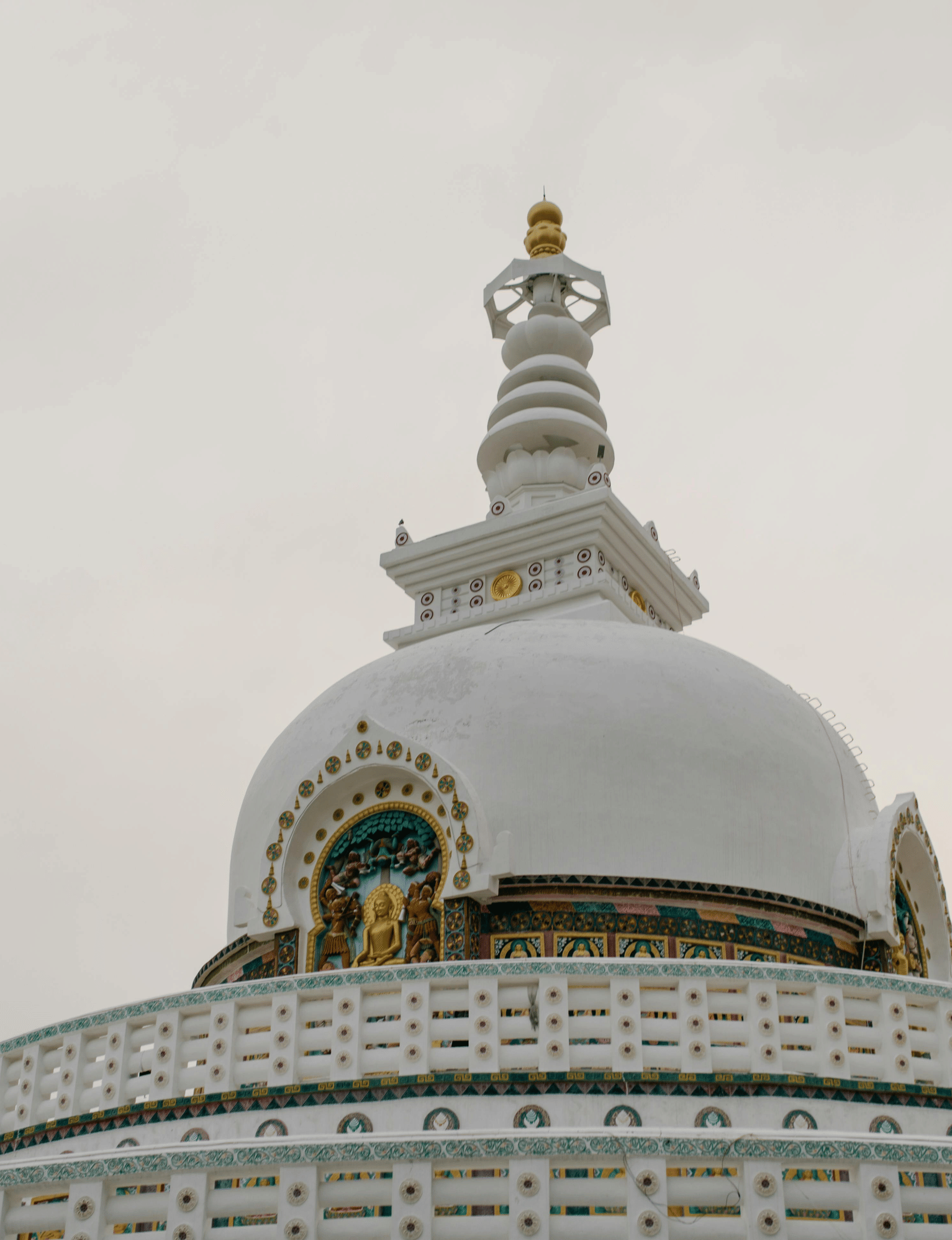 The upper part of a large, white Buddhist stupa with detailed gold trim and a tiered spire against a pale sky.