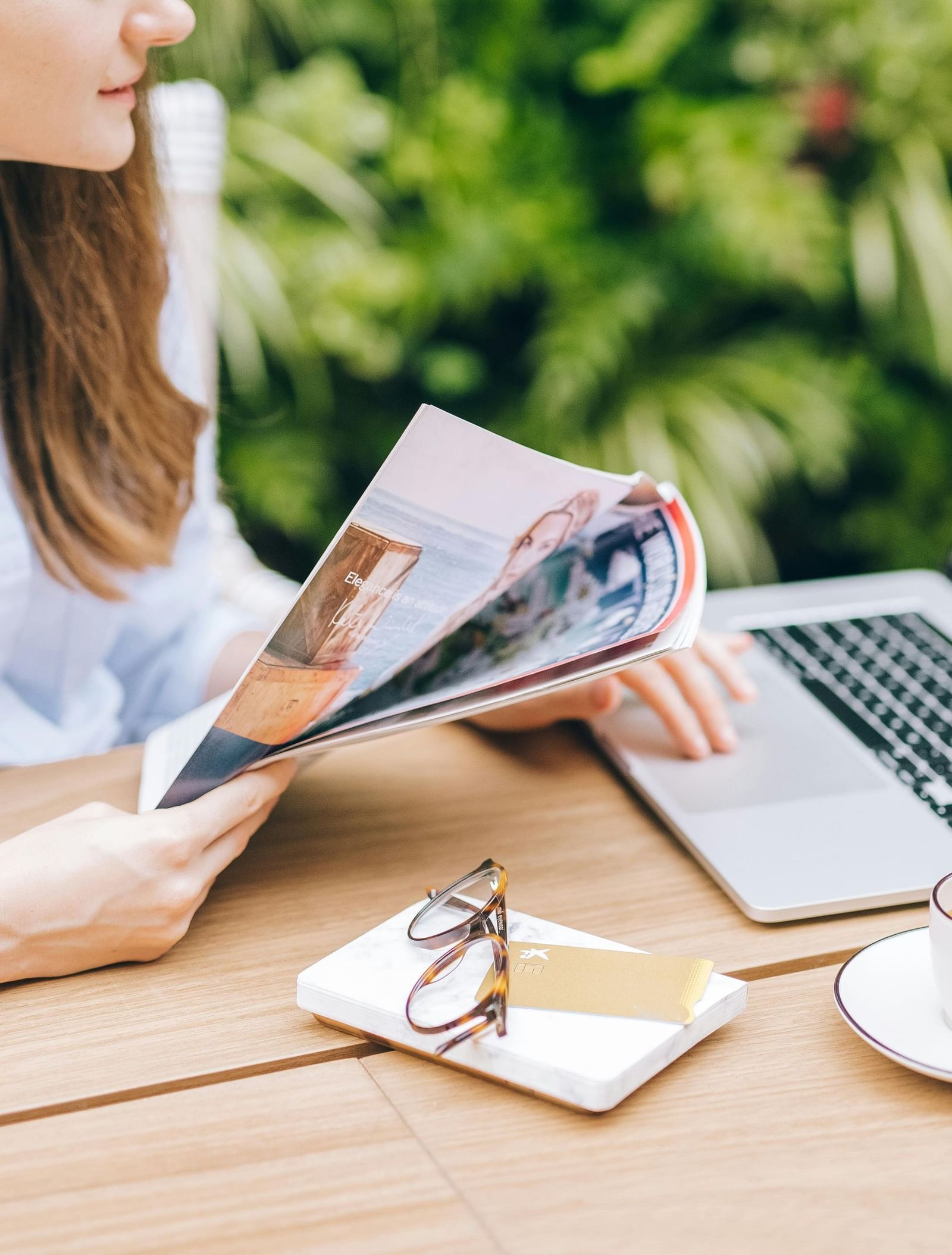 A person with long hair sitting at an outdoor table with a laptop, a cup of coffee, and a magazine.