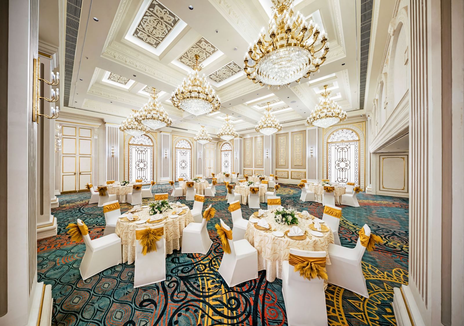 A grand hall prepared for a reception, showcasing elegant round tables with golden accents and white chairs, all beneath a ceiling adorned with multiple sparkling chandeliers.