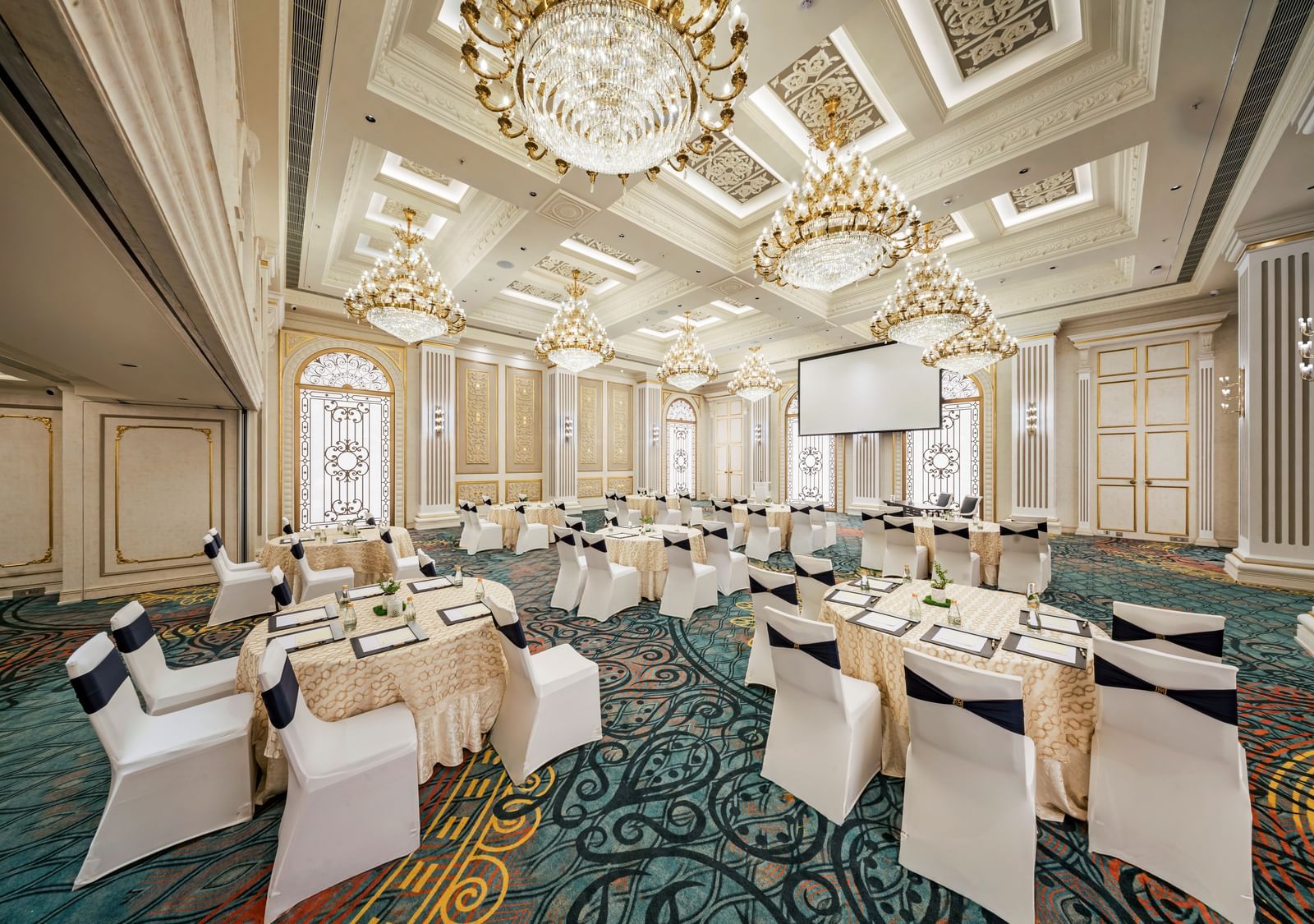A grand hall prepared for a reception, showcasing elegant round tables with golden accents and white chairs, all beneath a ceiling adorned with multiple sparkling chandeliers.