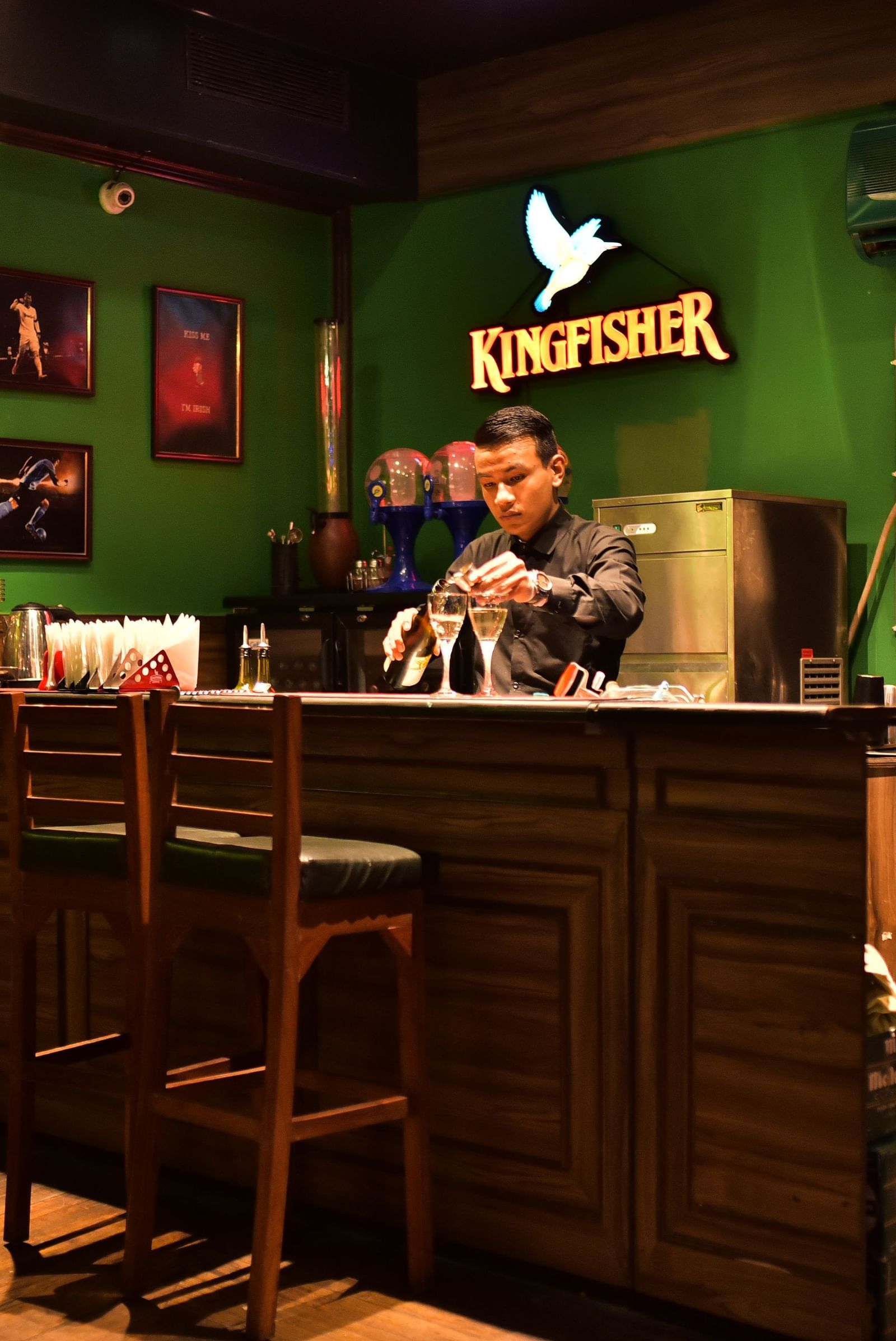 A person mixing a drink behind a bar with a green wall and a wooden counter at the Polo Hotel Shillong.