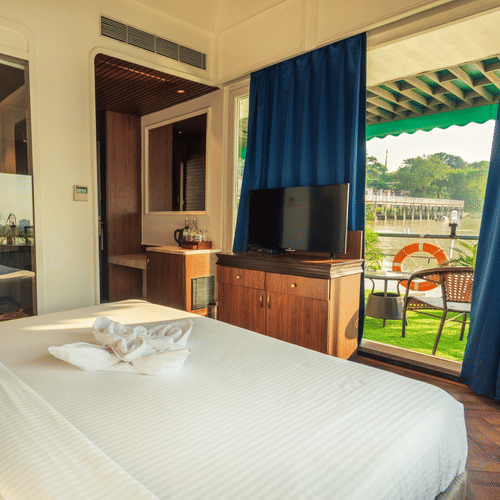 A bright bedroom with direct access to a balcony overlooking a water body, equipped with a large bed, TV, and dark wood furniture.