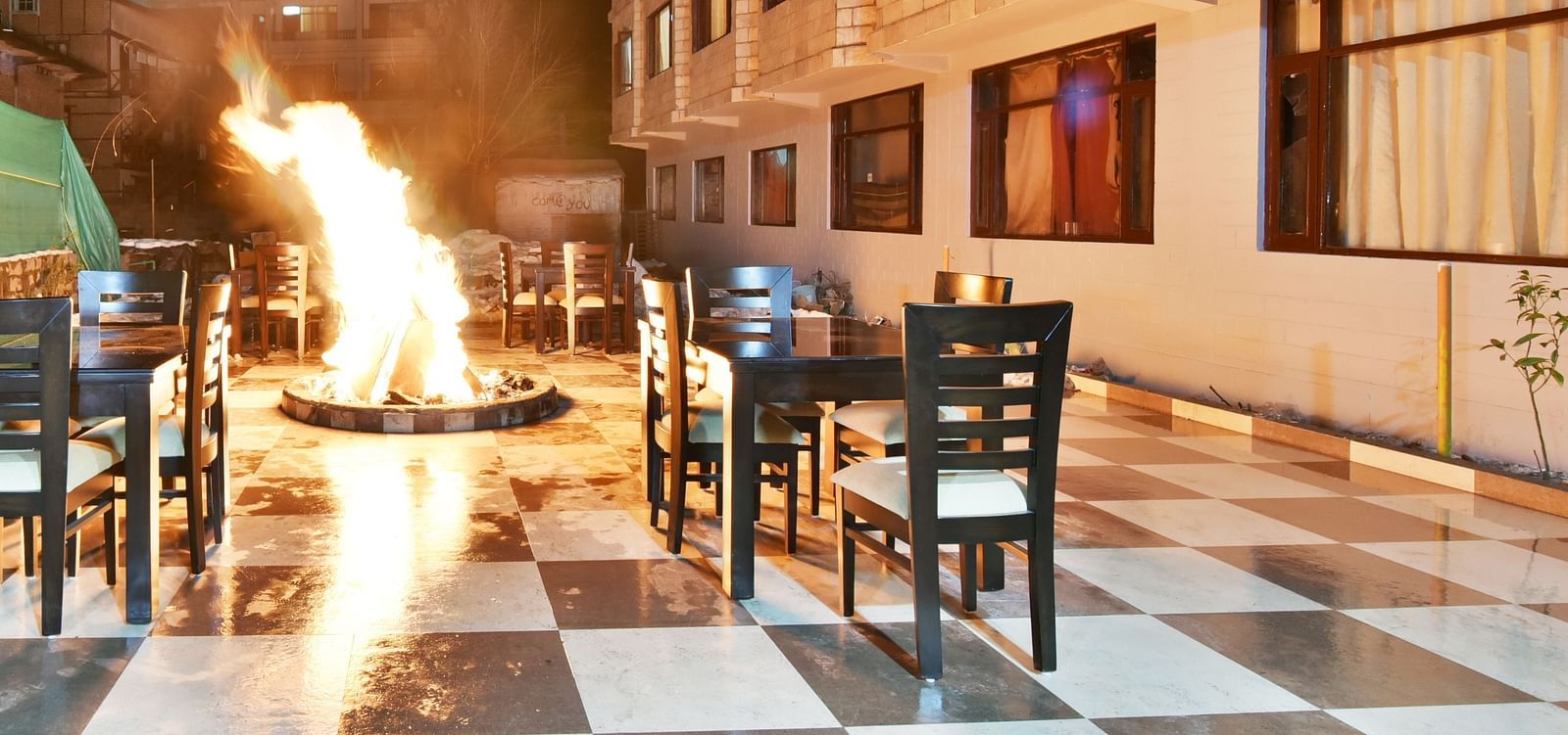 The image shows an outdoor dining area at night with a large bonfire in the centre, surrounded by tables and chairs | DLS Kapoor Resort Manali