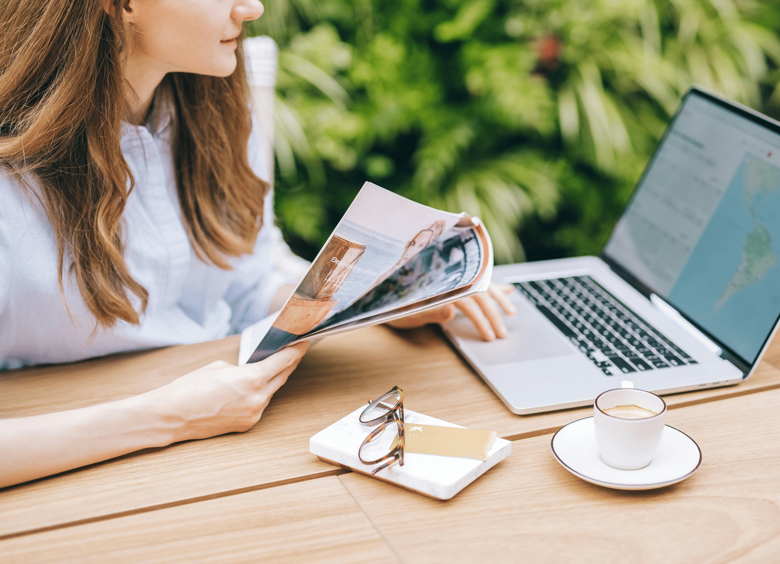 A person with long hair sitting at an outdoor table with a laptop, a cup of coffee, and a magazine.