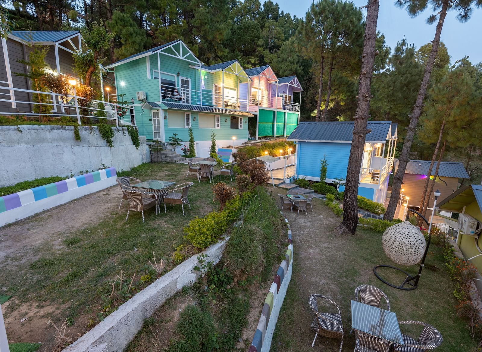 A scenic hillside resort with colourful cottages surrounded by tall pine trees, outdoor seating areas with tables and chairs, and landscaped garden paths at dusk - DLS Nature Trinket Resort, Dalhousie.