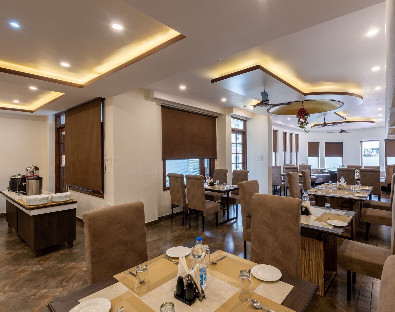 A restaurant with beige seating, tables set with plates, glasses, and cutlery. The space features modern lighting, brown window blinds, and a buffet area - DLS Meghavan Resort, Dharamshala