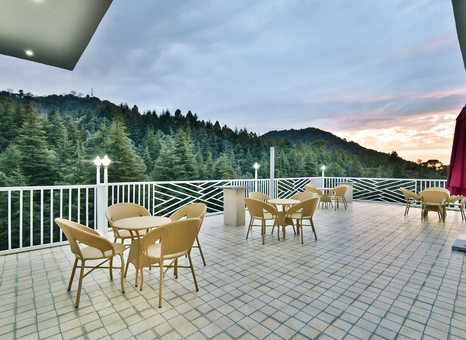 Open terrace area with chairs around tables with a background of trees at DLS Ark Holiday Inn, Banikhet at evening.