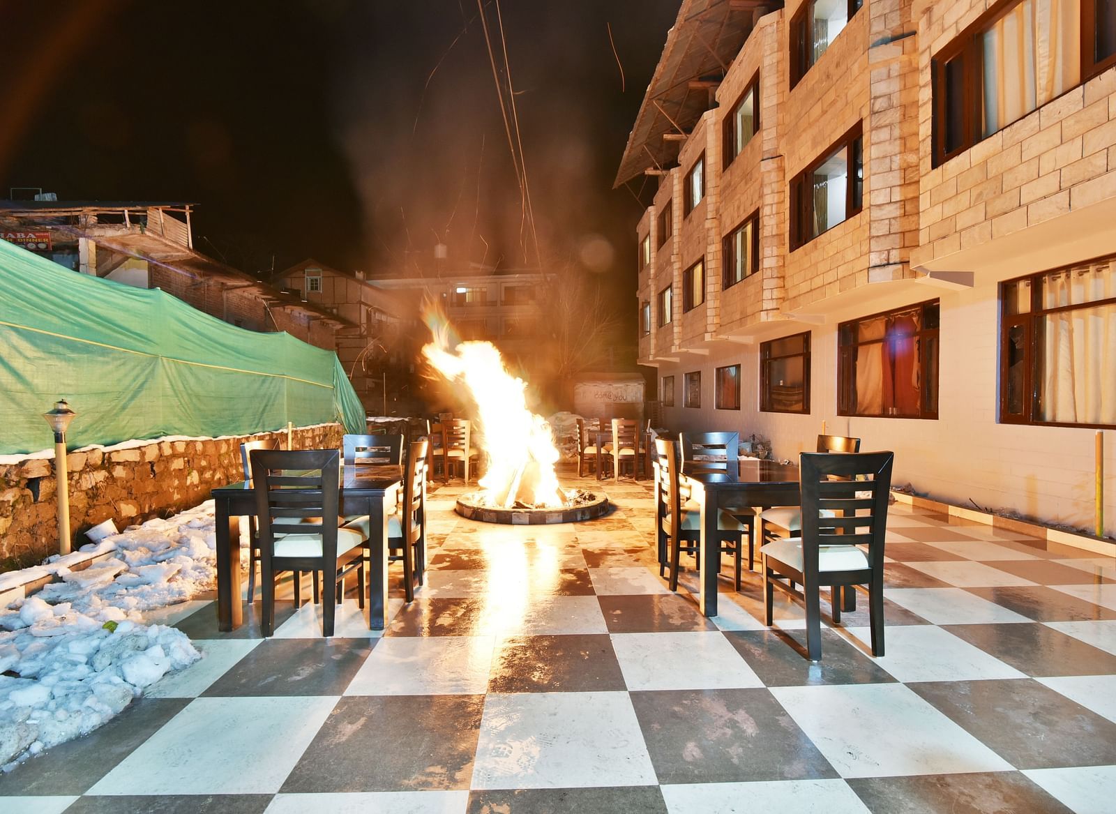 The image shows an outdoor dining area at night with a large bonfire in the centre, surrounded by tables and chairs | DLS Kapoor Resort Manali