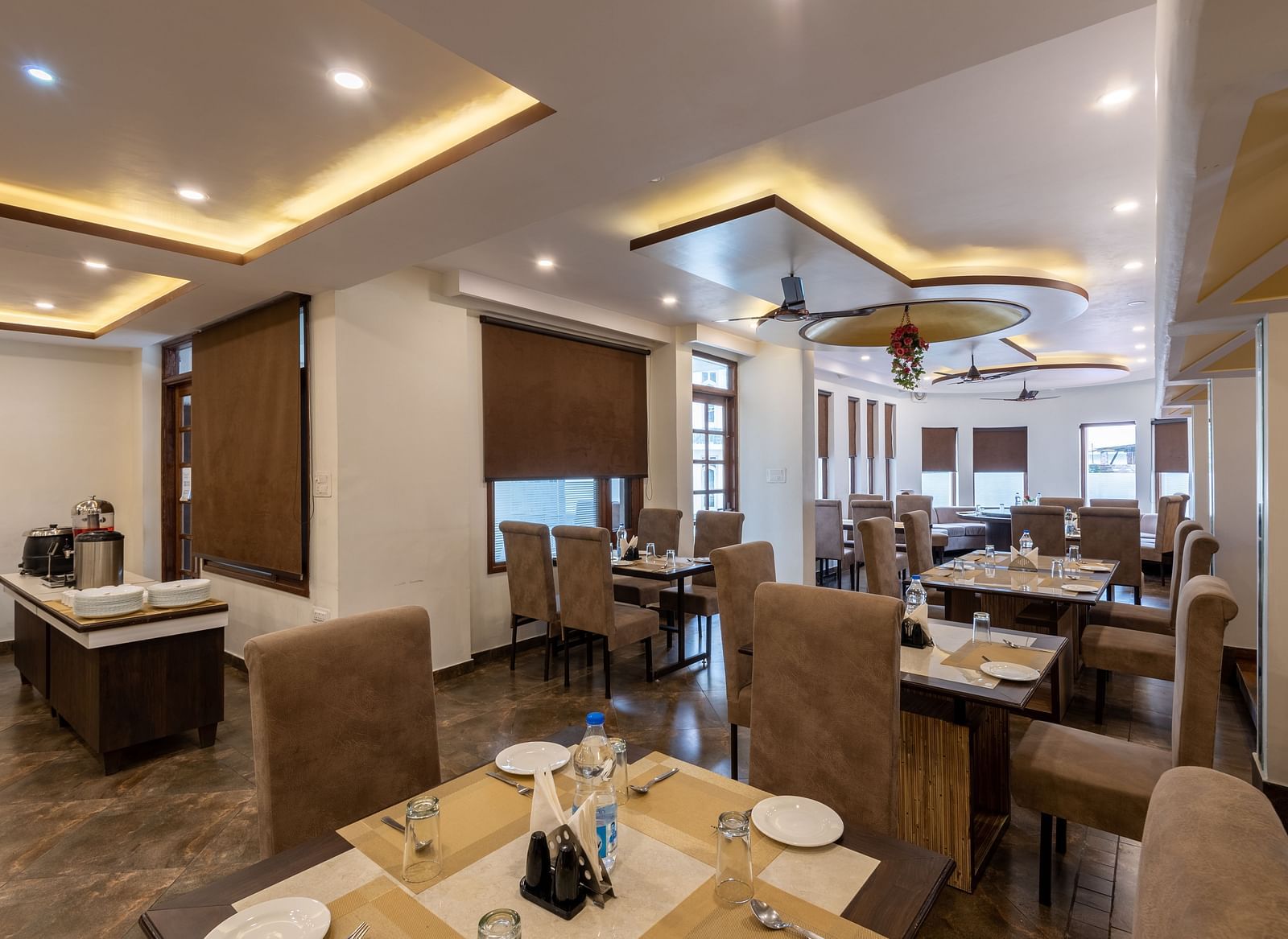 A restaurant with beige seating, tables set with plates, glasses, and cutlery. The space features modern lighting, brown window blinds, and a buffet area - DLS Meghavan Resort, Dharamshala