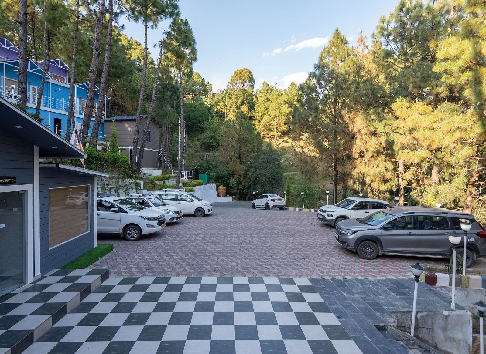 View of a paved parking lot with multiple cars parked, bordered by a forest of tall pine trees and colourful buildings, with a black-and-white checkered tile area in the foreground - DLS Nature Trinket Resort, Dalhousie.