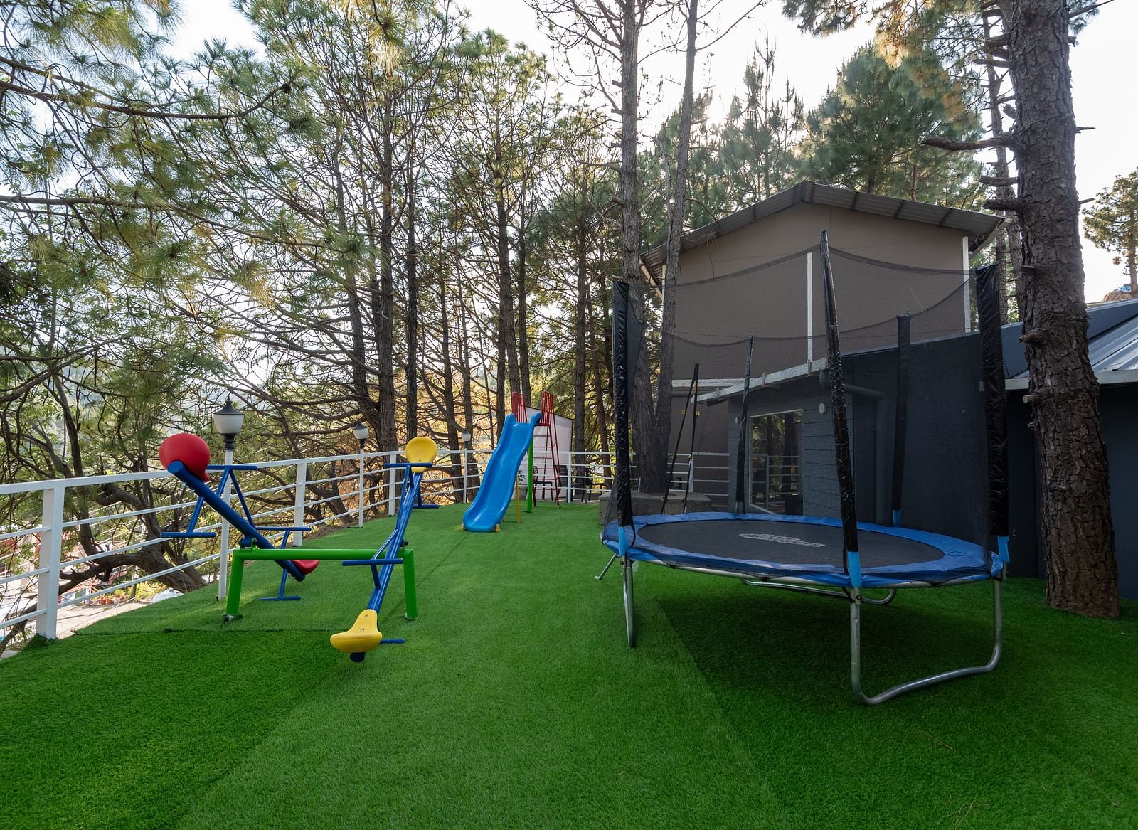 A children's play area on green artificial turf with a blue trampoline, a blue slide, and a colourful seesaw, surrounded by tall pine trees and a safety railing, next to a wooden structure - DLS Nature Trinket Resort, Dalhousie.