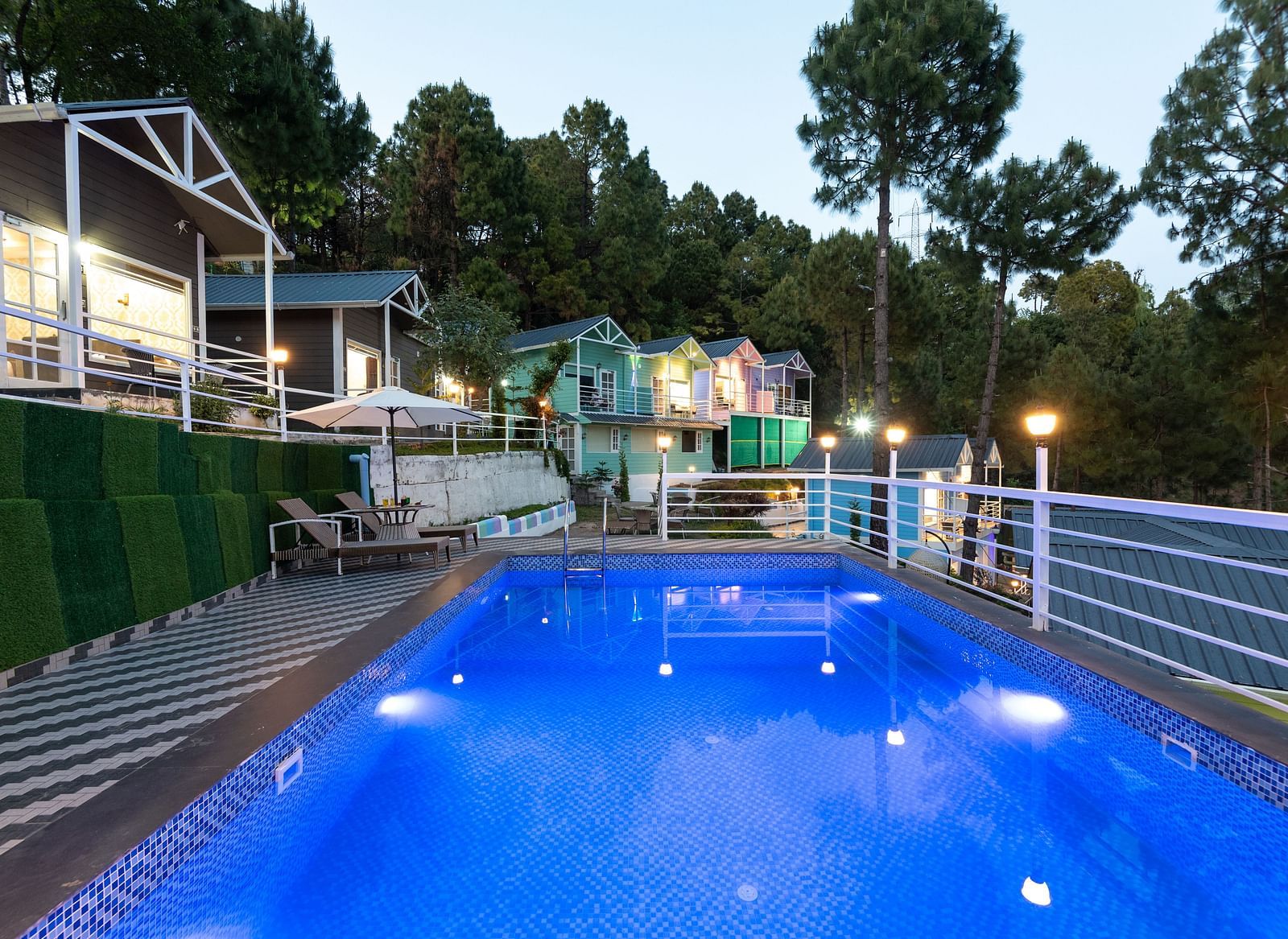 Tranquil evening view of a lit swimming pool surrounded by safety railings and striped flooring, with cosy hillside cottages and tall pine trees in the background - DLS Nature Trinket Resort, Dalhousie.