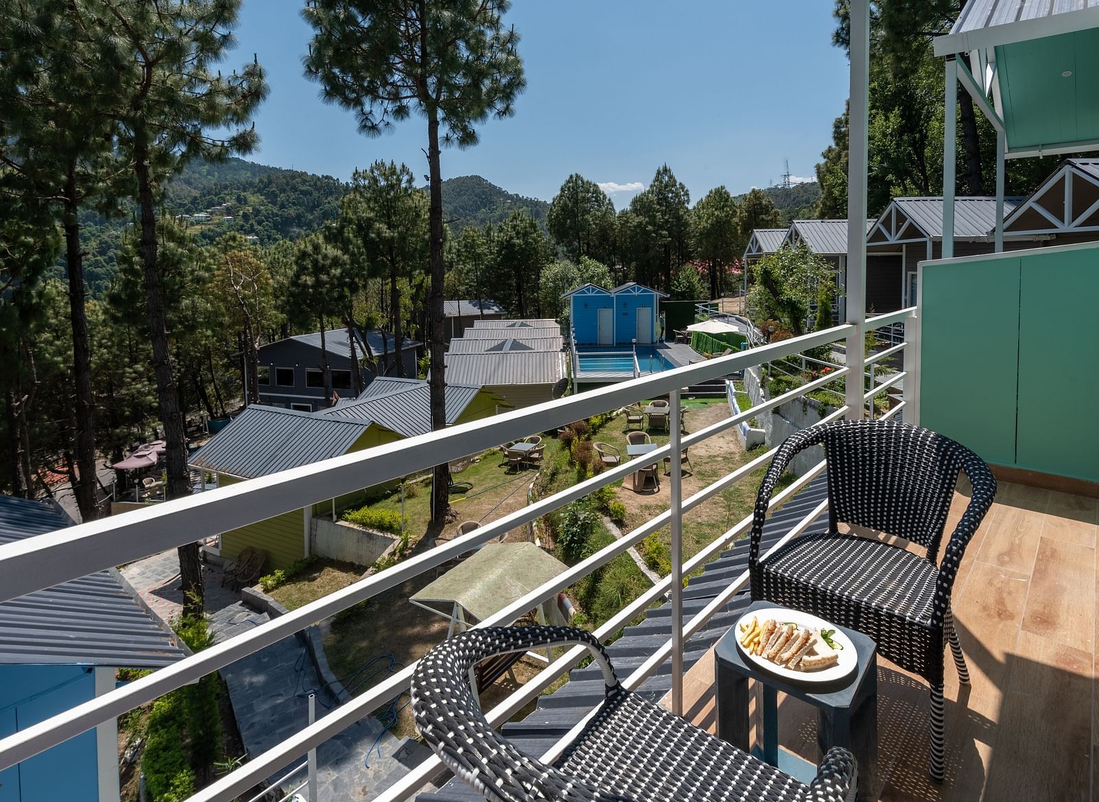 A balcony at DLS Nature Trinket Resort, Dalhousie, features two black wicker chairs and a small table with a plate of food, overlooking a scenic view of buildings, greenery, and tall trees under a clear sky.