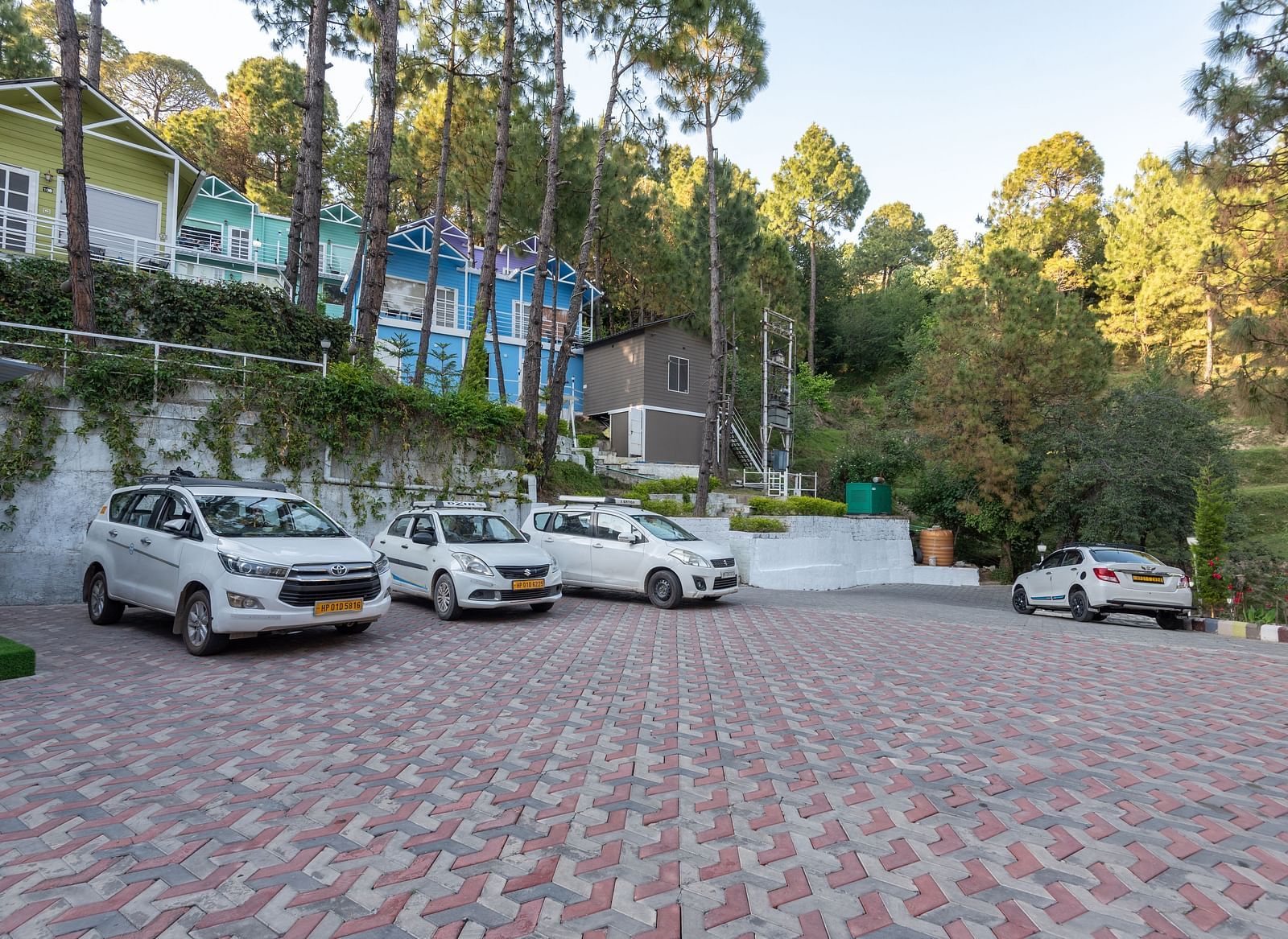  Spacious parking area with several white cars parked on a tiled surface, set against a backdrop of colourful hillside cottages and tall pine trees - DLS Nature Trinket Resort, Dalhousie.