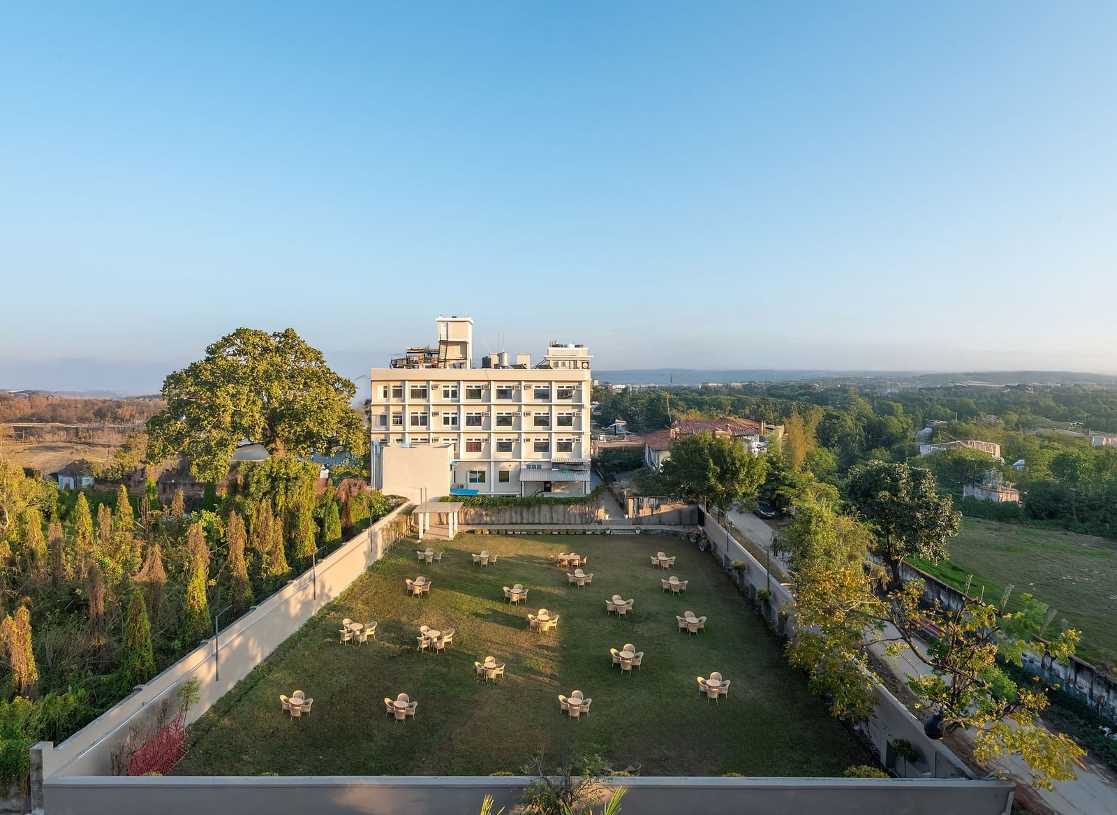  Lawn area at DLS Park Grace, Haridwar featuring several sets of tables and chairs surrounded by luscious green trees and site of the clear blue sky.