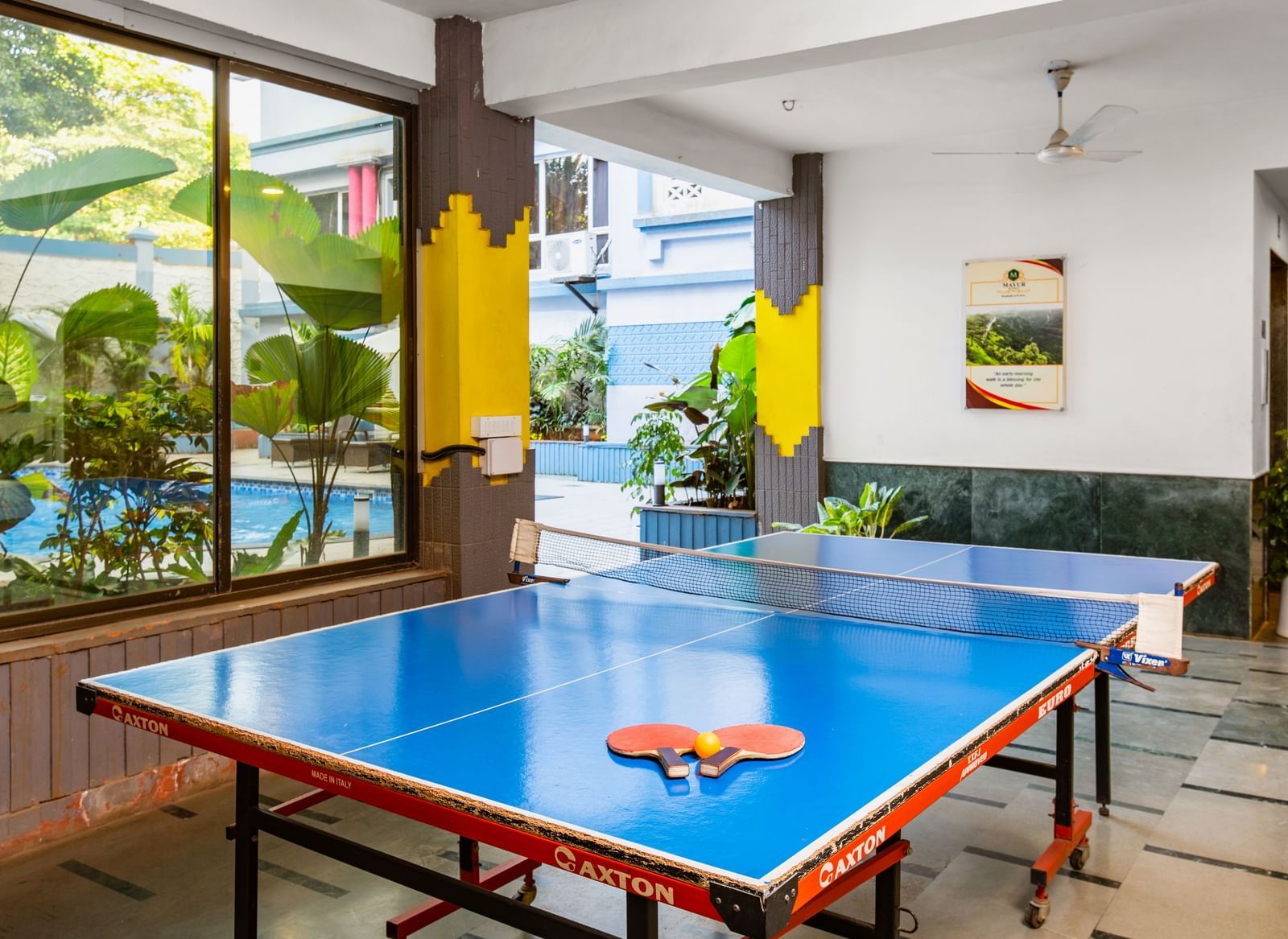 Indoor recreational room at Mayur Retreat featuring a table tennis setup, potted plants, and glass walls offering a mix of fun and relaxation in a lively environment.