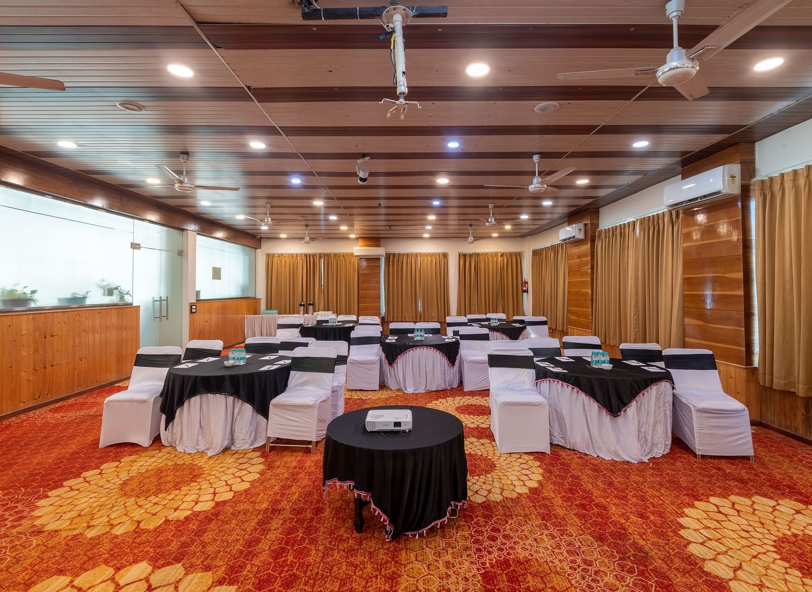 Well lit banquet hall at Regenta Inn DLS, Rishikesh with circular tables and chairs surrounding them.