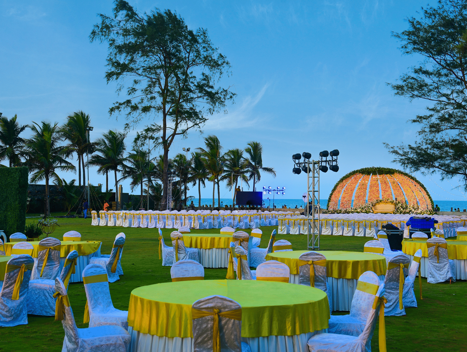 An outdoor evening event setup on a green lawn with round tables draped in yellow, featuring a lit stage and palm trees against a blue sky - Grande Bay Resort & Spa, Mamallapuram