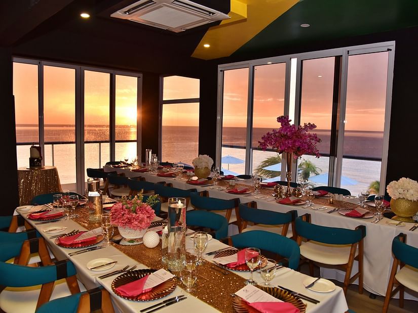 An interior view of a dining area at S Hotel Montego Bay with a long table set for a meal, teal and wooden chairs, and large windows showing a sunset over the ocean.