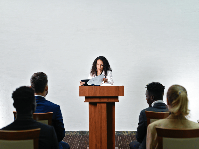 A woman standing behind a wooden podium, reviewing documents while four people sit facing her, listening attentively. The background is a plain white wall, and the audience members are dressed in formal attire.