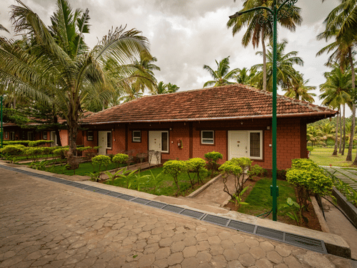 Facade of the River View Suite with gardens, a pathway, and coconut trees under a cloudy sky at Ibex River Resort, Pollachi, one of the best hotels near Aliyar Dam.
